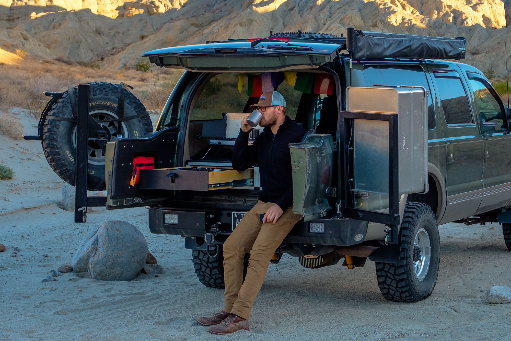 Chris Cordes sits on the back of an SUV with the trunk open, drinking from a mug in a desert setting. Camping gear and storage boxes are organized in the back of the vehicle. Rocky hills are visible in the background.