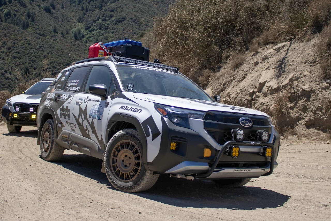 Two white Subaru rally cars with Leitner Designs off-road equipment parked on a dirt trail.