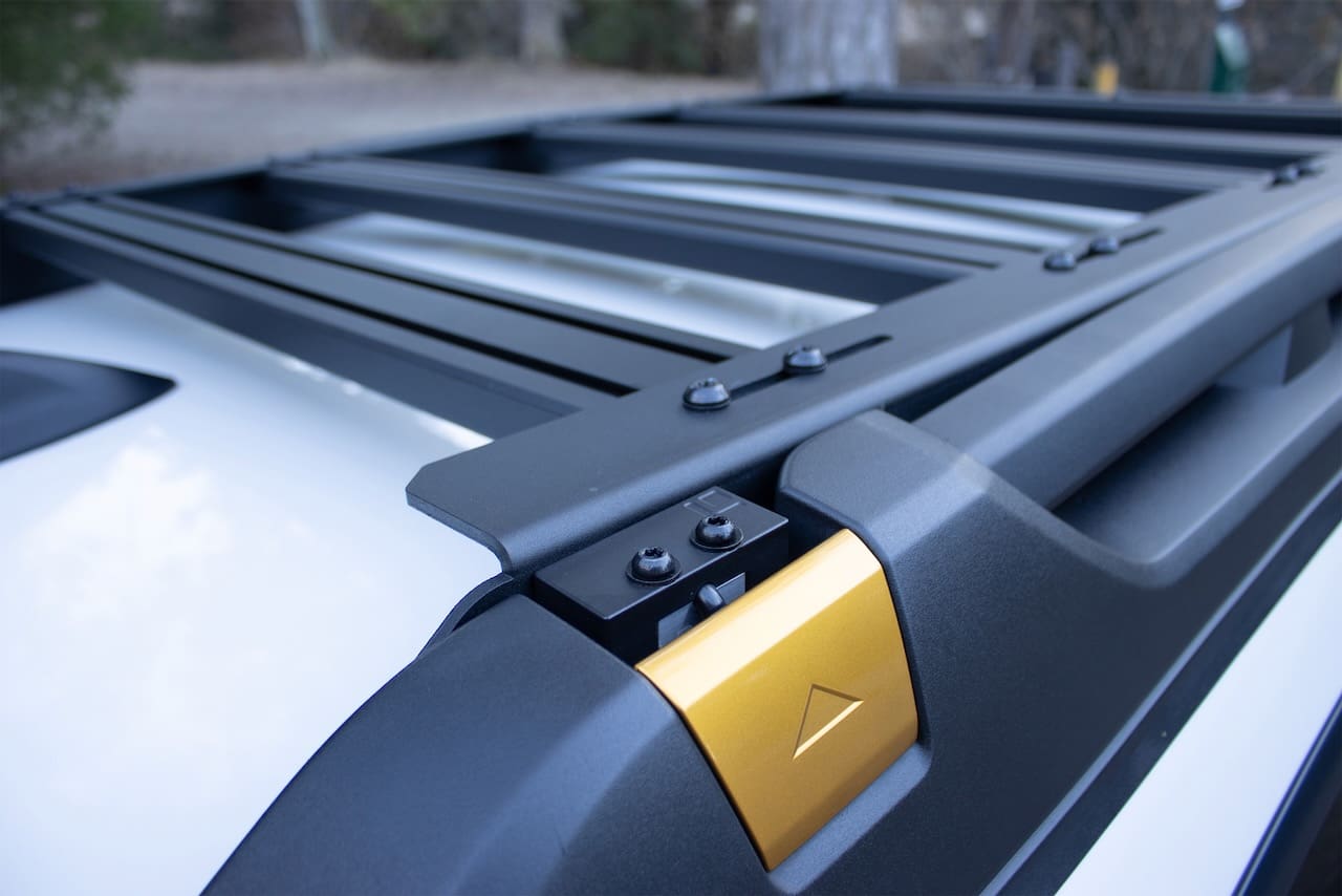 Black Leitner Designs metal roof rack installed on a white vehicle with a gold accent piece featuring an upward-pointing triangle.