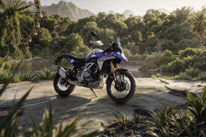 A white, blue, and red 2027 BMW F 450 GS adventure motorcycle parked on a stone outcrop overlooking a river with a tropical forest and a Ferris wheel in the background.