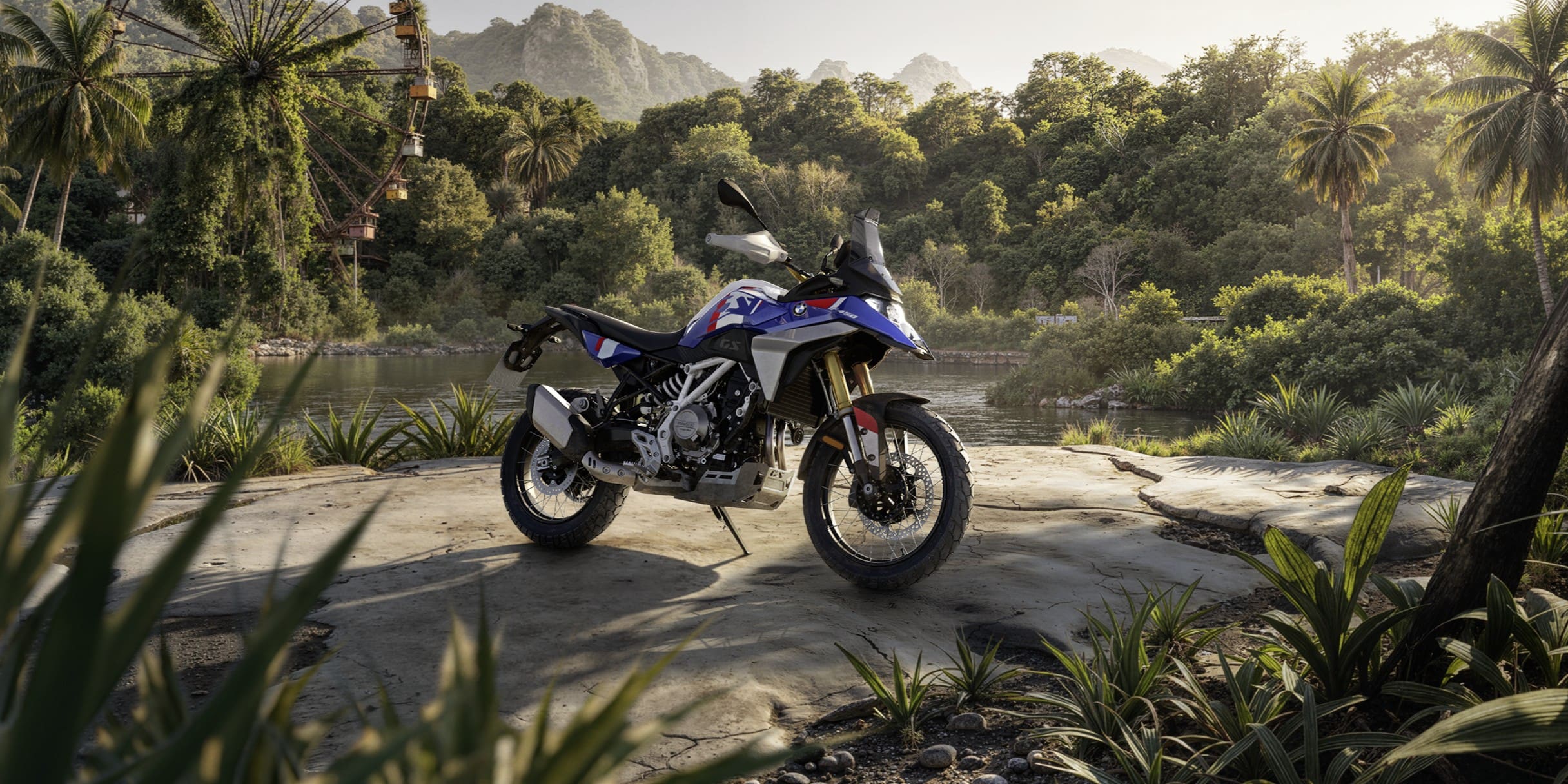 A white, blue, and red 2027 BMW F 450 GS adventure motorcycle parked on a stone outcrop overlooking a river with a tropical forest and a Ferris wheel in the background.