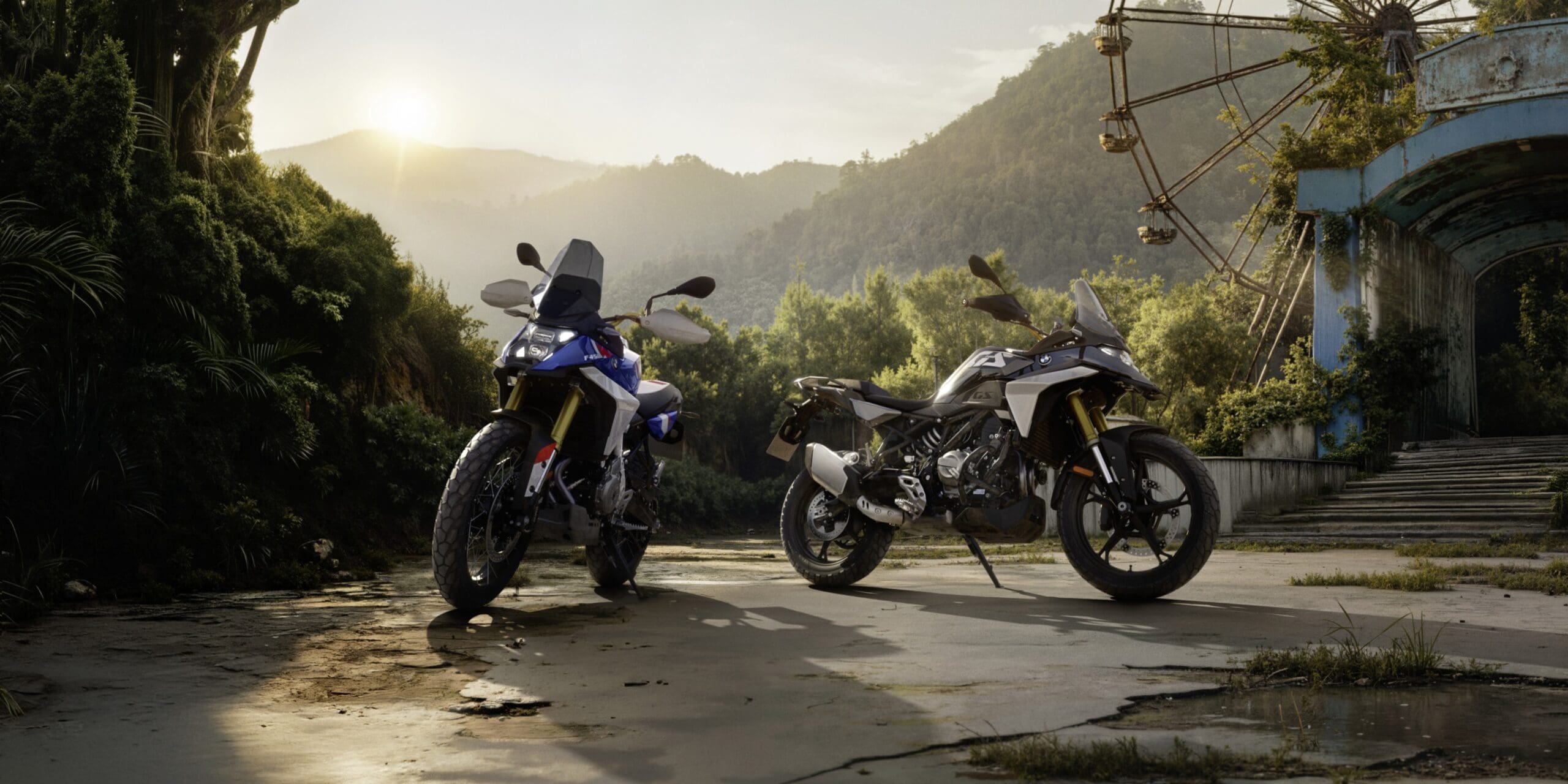 Two BMW Motorrad adventure motorcycles parked on a paved road near a mountain landscape and an old, rusted Ferris wheel.