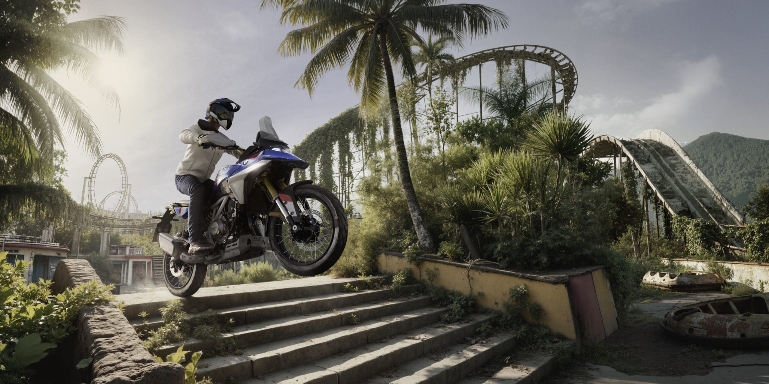 A motorcyclist rides a blue and white BMW Motorrad adventure bike up a set of stone stairs in an overgrown, abandoned amusement park.