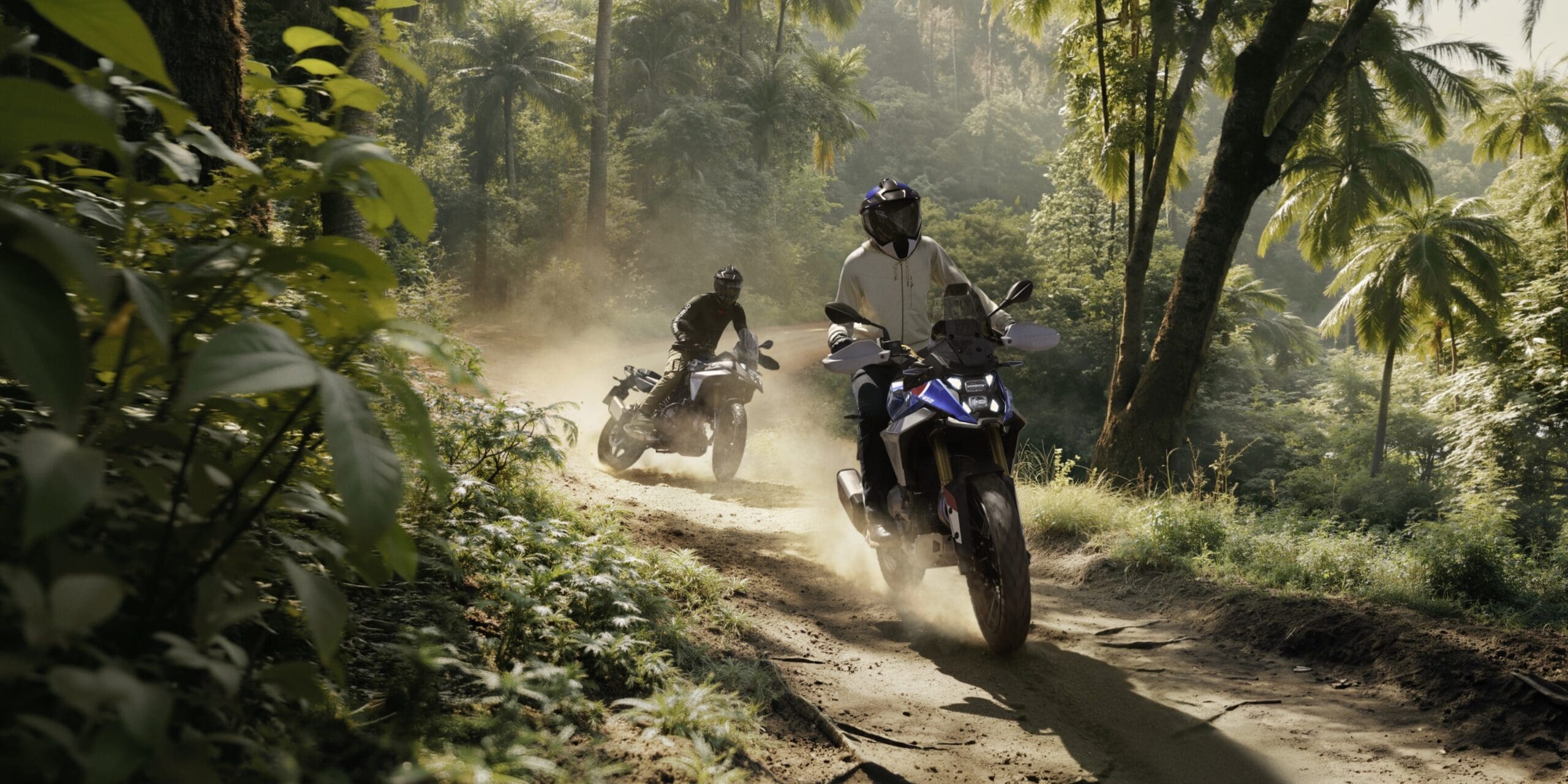 Two motorcyclists ride BMW Motorrad dual-sport bikes along a dusty, sunlit dirt path through a forest.