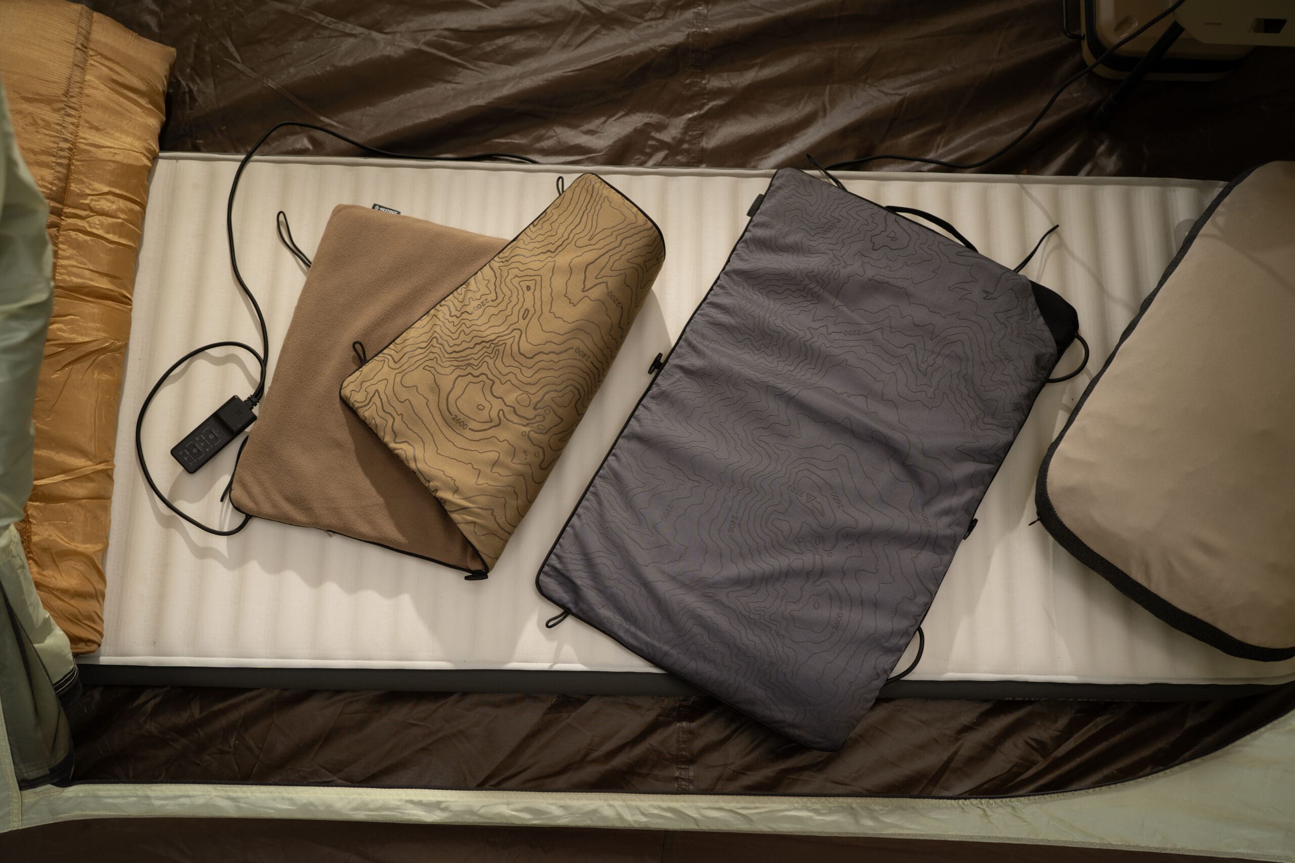Three rectangular heated seat pads with topographic map patterns resting on a white sleeping mat inside an overland industry tent.