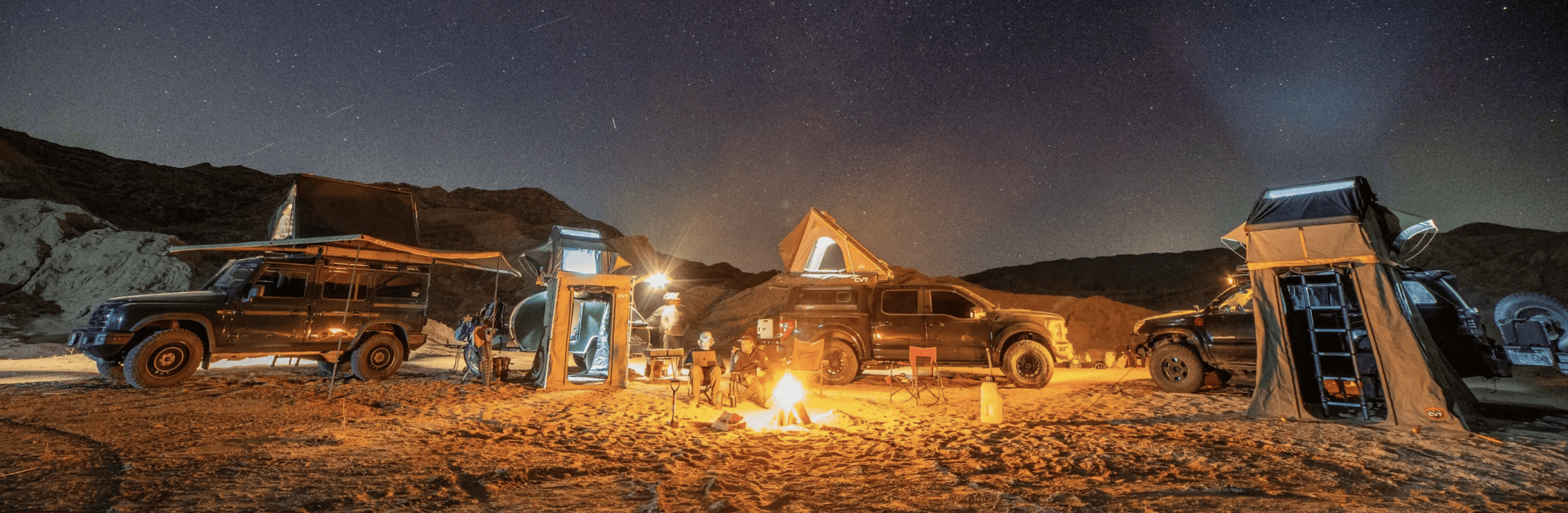 Three off-road vehicles with rooftop tents parked in a desert at night for adventure travel, with two people sitting around a campfire.