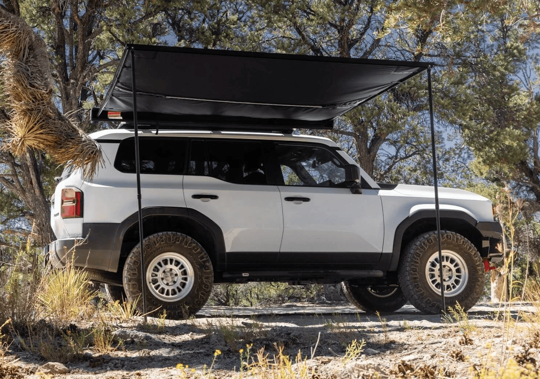 A white SUV with a deployed side awning parked in a wooded area, perfect for adventure travel.