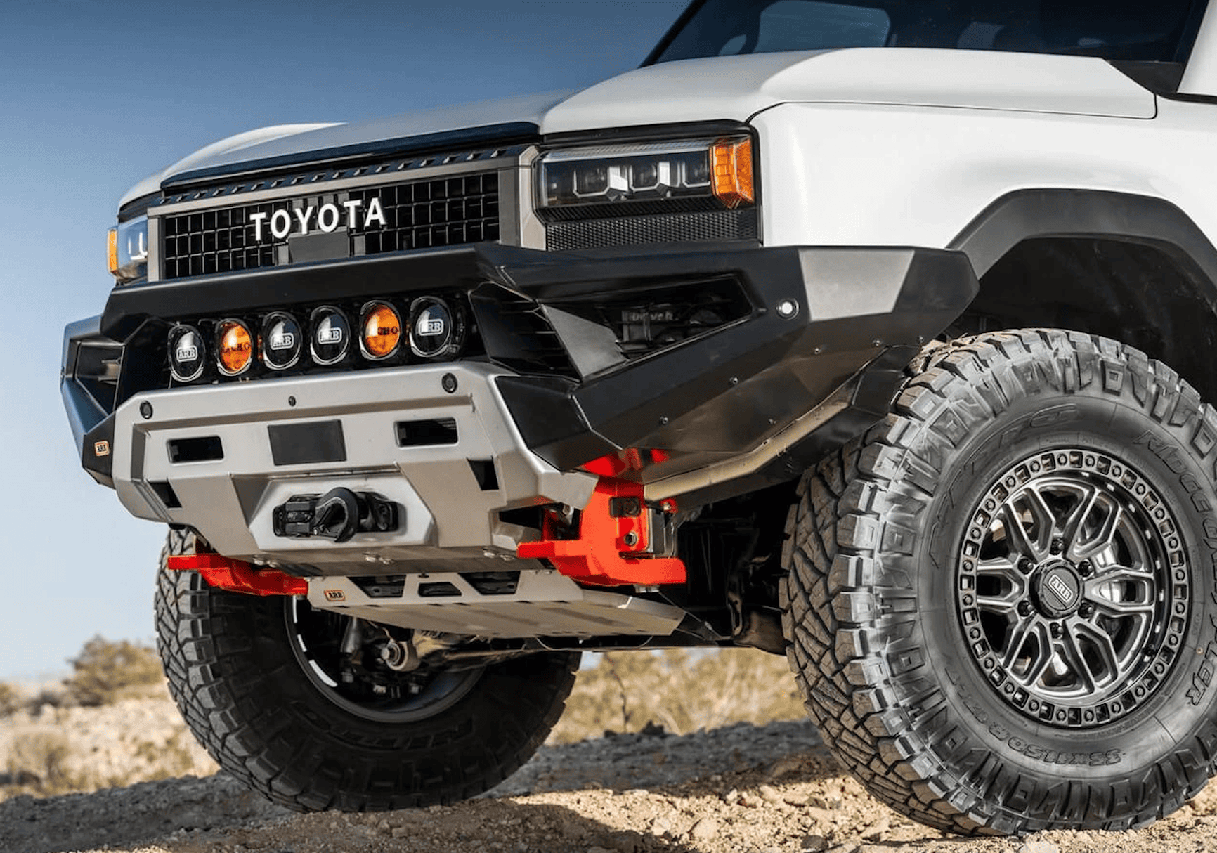 Front view of a white Toyota off-road truck for overland news, featuring an aftermarket ARB bumper, integrated circular lights, and red recovery hooks.