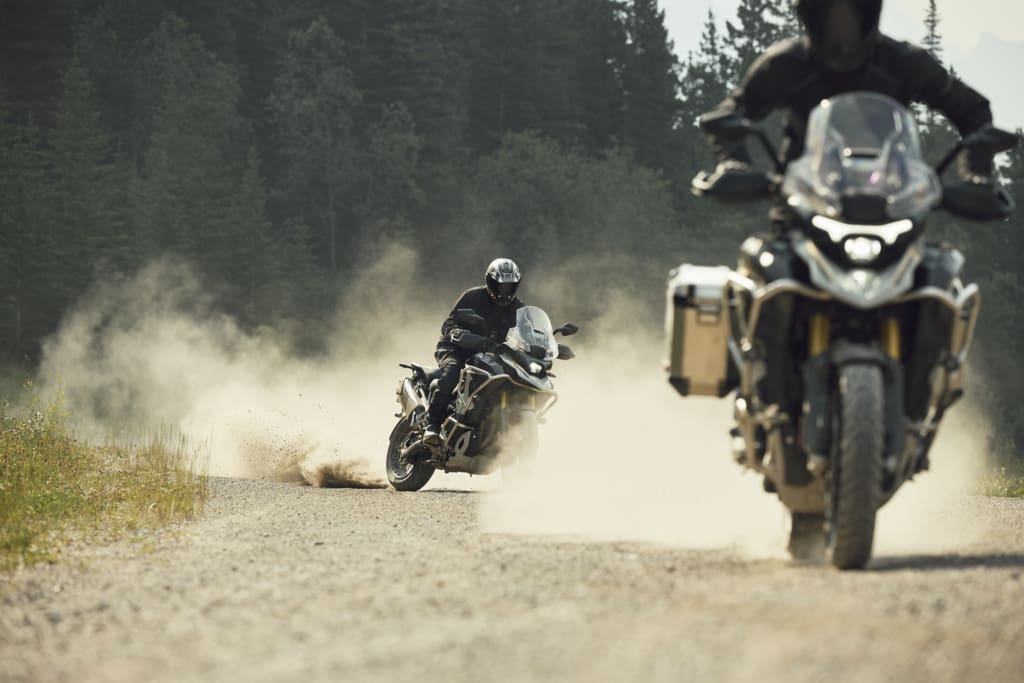 Two motorcyclists riding Triumph Tiger 1200 adventure bikes on a dusty gravel road.