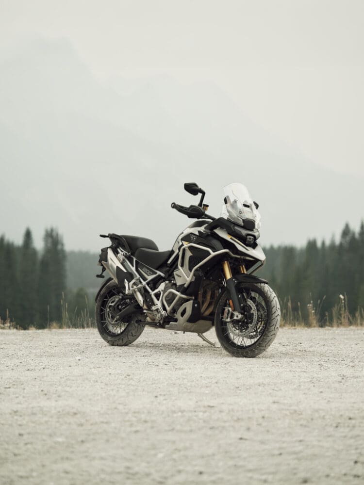 Triumph Tiger 1200 adventure motorcycle parked on a gravel surface against a backdrop of trees and misty mountains, ready for any necessary motorcycle upgrades.