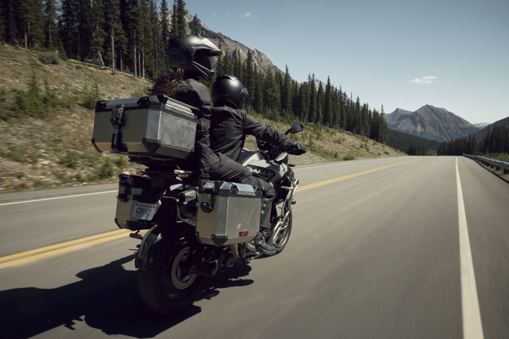 Two people wearing helmets ride a Triumph Tiger 1200 equipped with storage cases along a mountain road.