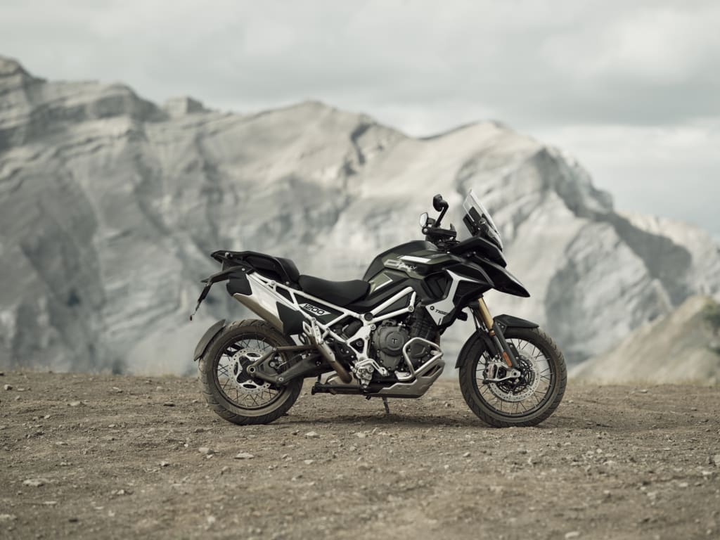 A dark green Triumph Tiger 1200 adventure motorcycle parked on a dirt path with large mountains in the background, showcasing the capabilities of modern ADV bikes.