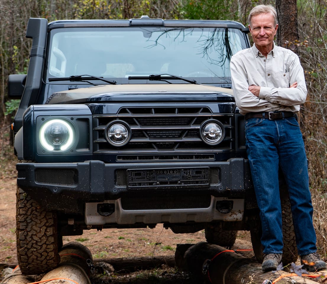 Jonathan Hanson stands with his arms crossed next to a Land Cruiser parked on logs in a wooded area.