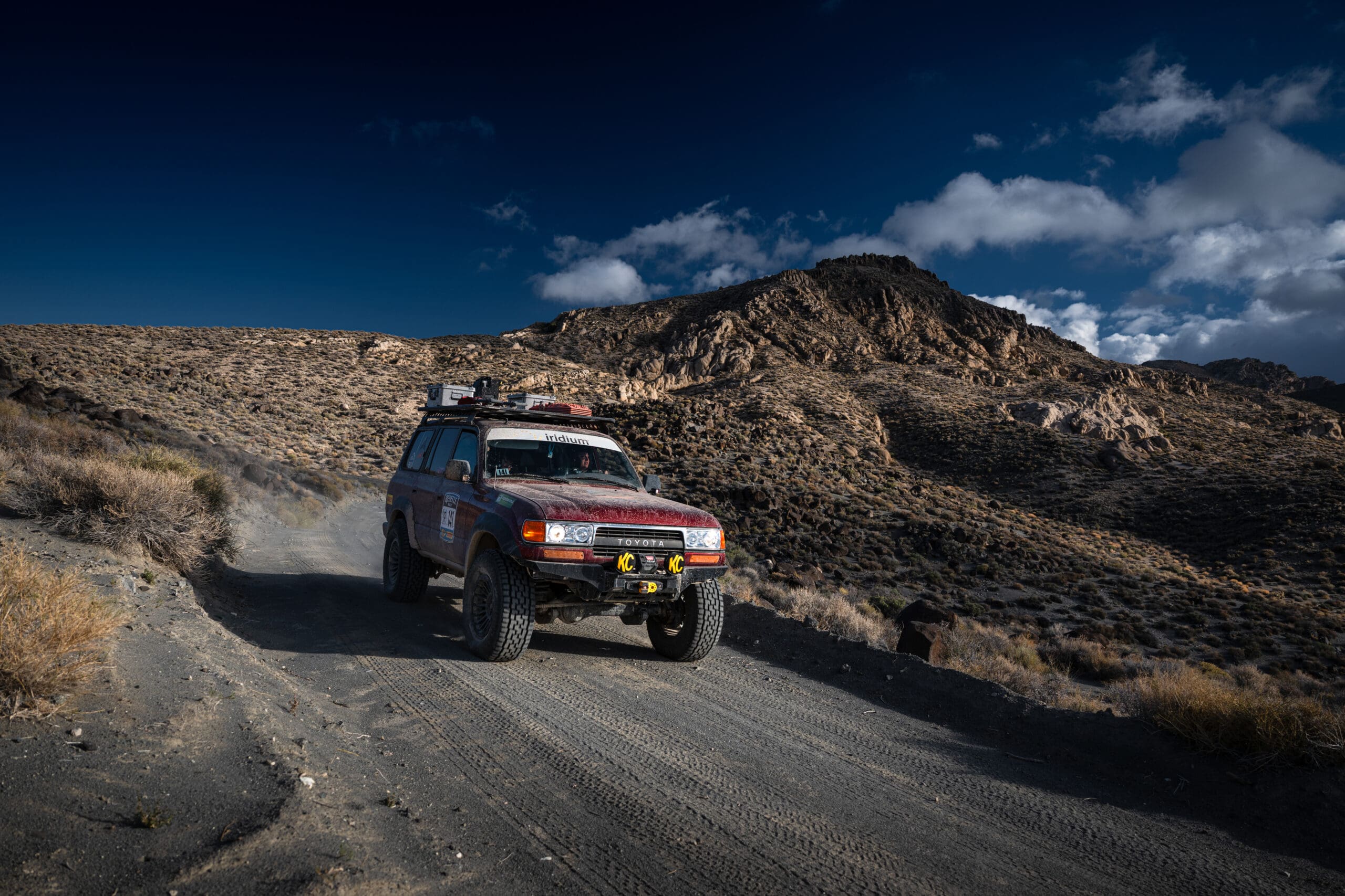 A red Toyota Land Cruiser driving on a dirt road in a rocky, mountainous landscape, captured for Overland News.