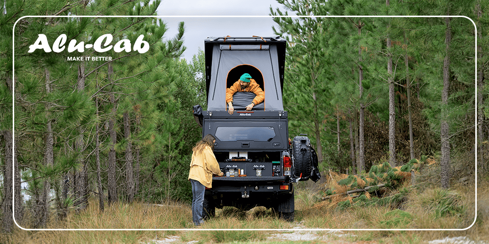 A person stands at the rear of an off-road vehicle with a rooftop tent as another sits inside, a scene captured for Overland Weekly in the pine forest.