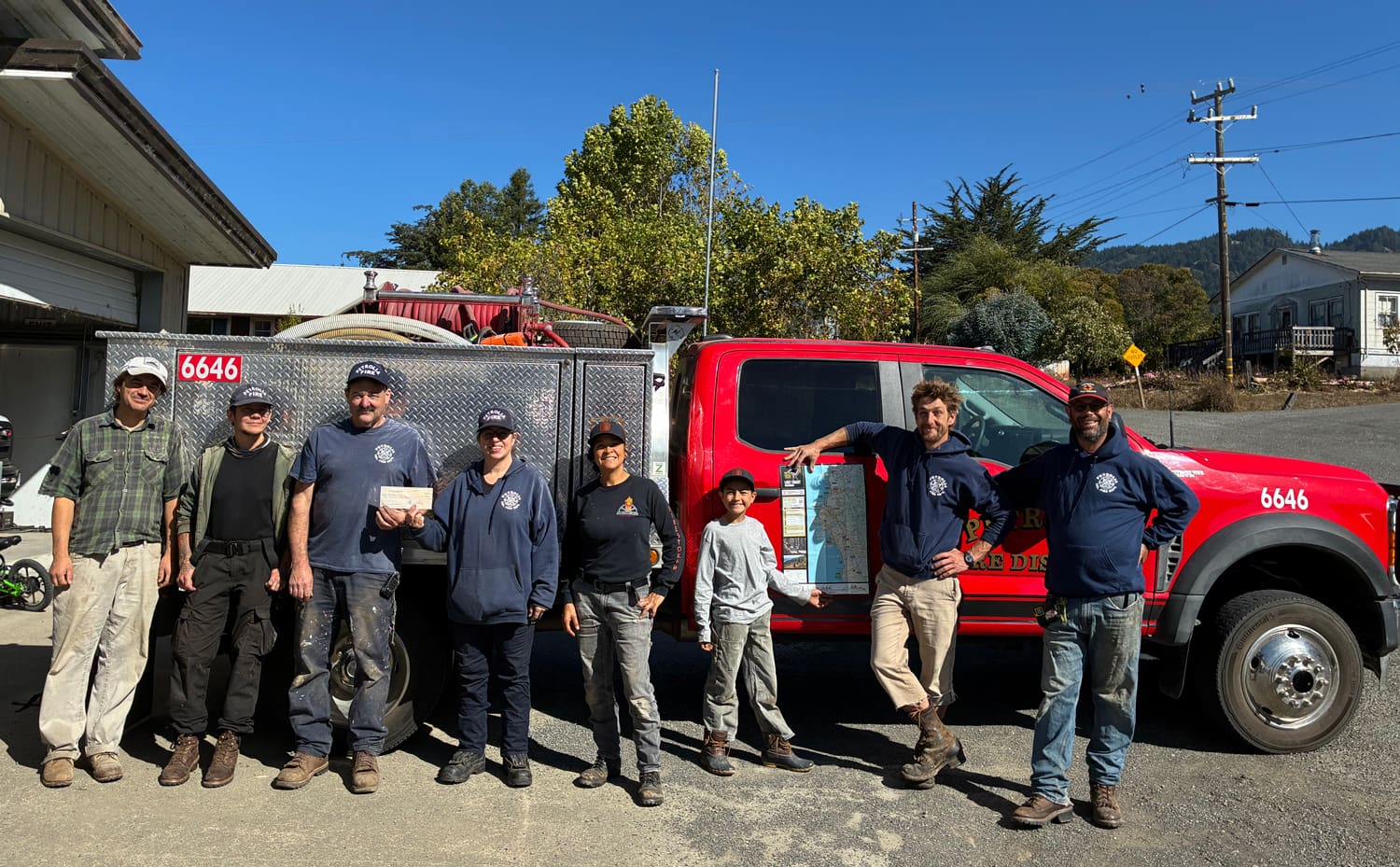 Seven people stand before a red fire truck for Overland News, with one person holding a check and another holding a map.