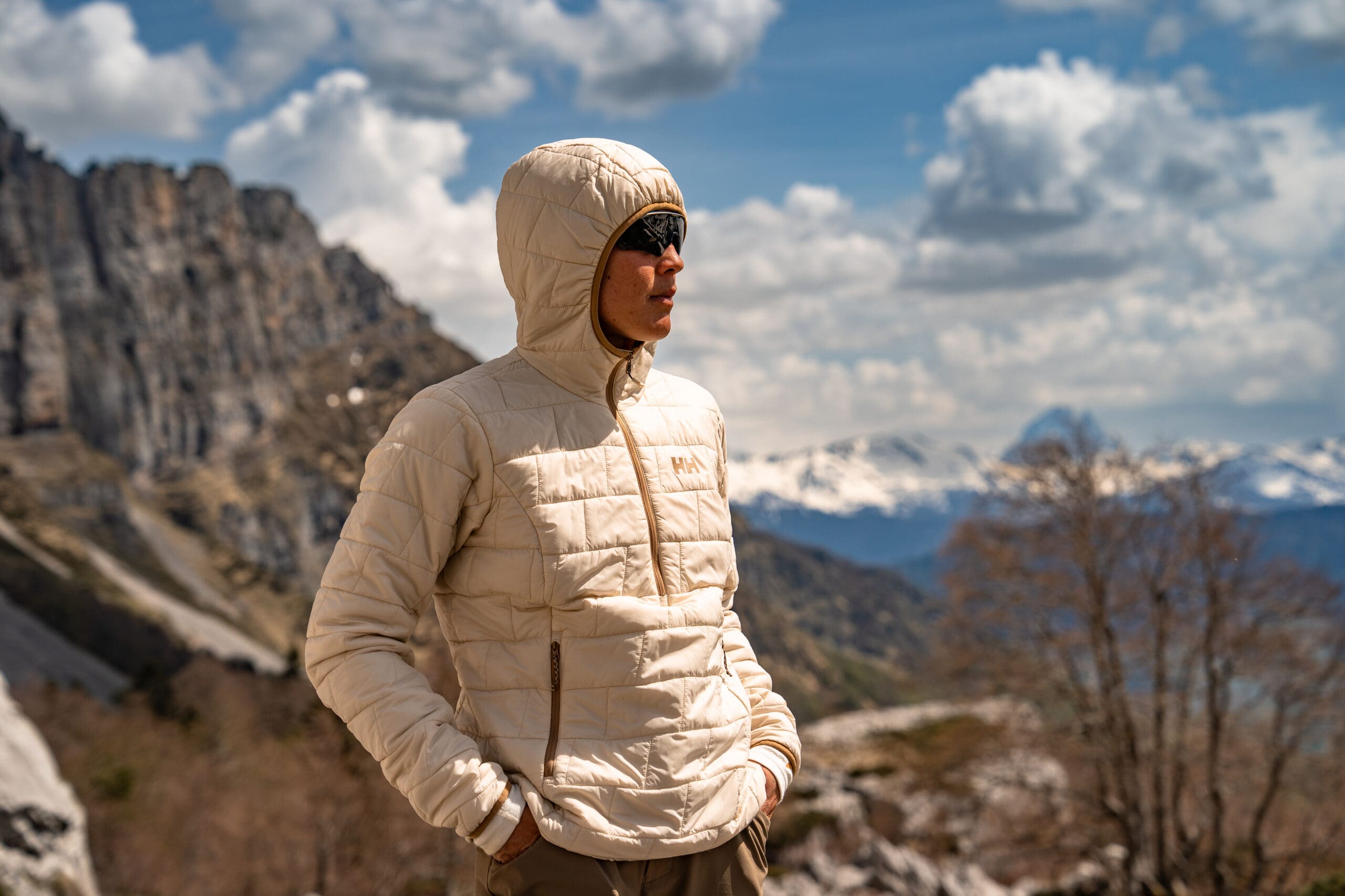 A person wearing a hooded beige quilted jacket and sunglasses stands on a mountain overlook, capturing the latest overland news.