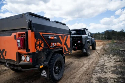 A black and orange Patriot Campers off-road trailer being towed by a black pickup truck on a dirt track.