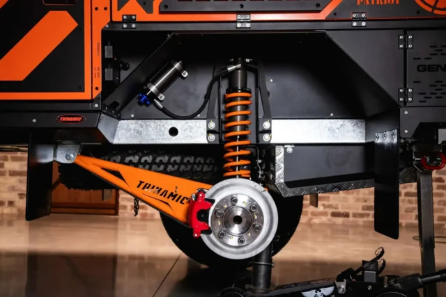 Close-up of a Patriot Campers orange and black vehicle suspension system featuring a coil spring and a brake rotor branded with the Trinamic logo.