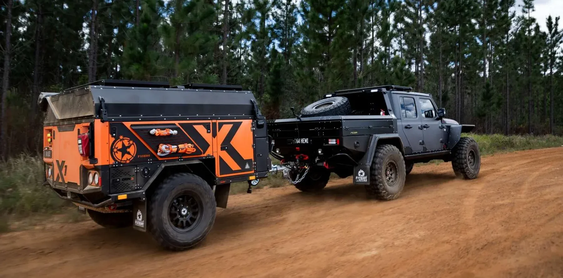 A black pickup truck towing an orange and black Patriot Campers Gen 3 X1 off-road trailer on a dirt path through a forest.