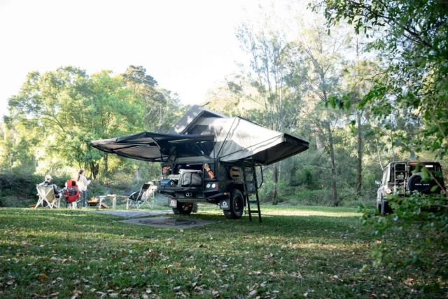 A rugged Patriot Campers off-road trailer with a deployed rooftop tent and awning parked in a grassy clearing with a campfire and a group of people nearby.