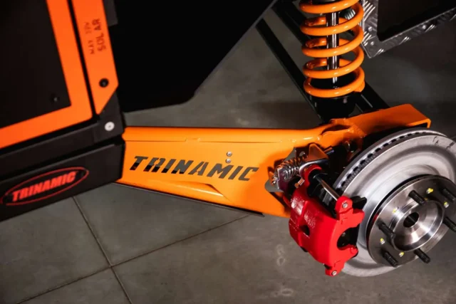 Close-up of a bright orange Patriot Campers suspension arm branded Trinamic, featuring a bright orange coil spring, a red brake caliper, and a silver brake disc.