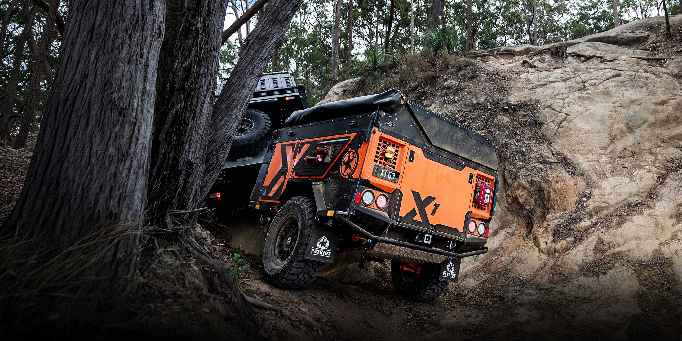 An orange Patriot Campers X1 trailer being towed by a vehicle off-road on a rocky, wooded trail.