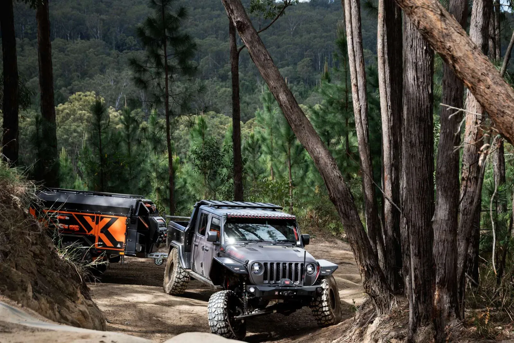 A gray Jeep Gladiator towing a black and orange Patriot Campers off-road trailer along a dirt path in a pine forest.