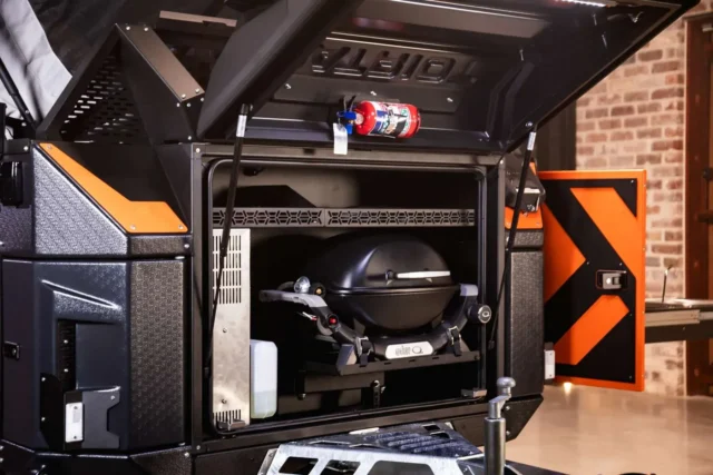 Open storage compartment on a black and orange Patriot Campers trailer containing a mounted Weber portable grill and a fire extinguisher attached to the inside of the hatch.