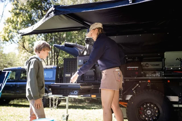 A woman and a young boy stand by an open Patriot Campers trailer in a grassy area.