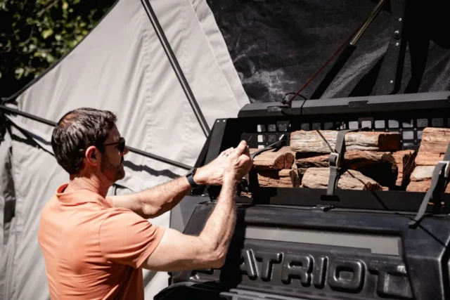 A man in an orange shirt secures a stack of firewood into a rack on the side of a Patriot camper.