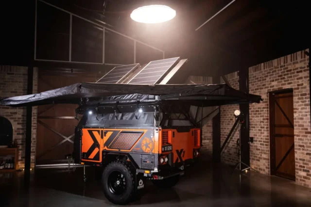 Orange and black Patriot Campers off-road trailer with a roof-mounted solar panel and extended awning parked in a brick garage.