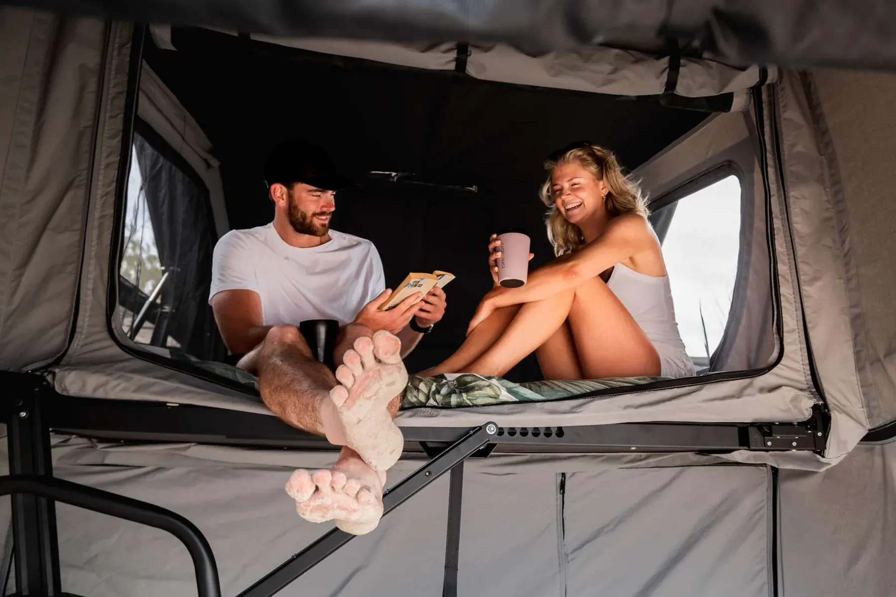 A man and a woman sitting inside a Patriot Campers rooftop tent, with the man reading a book and the woman holding a mug.
