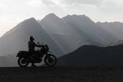 A motorcyclist in a helmet sits on a bike parked on a ridge, silhouetted against a mountain range at sunset, a scene typical of adventure travel.