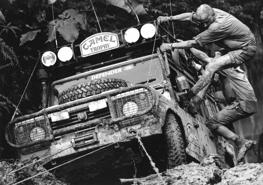 two men clinging to the outside of a muddy SUV